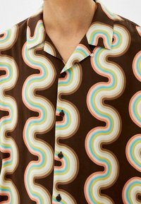 Korte mouwen shirt met een bruine basis, voorzien van golvende, meerkleurige patronen in roze, blauw en roomkleurig. Inclusief zwarte knopen en een kraag.