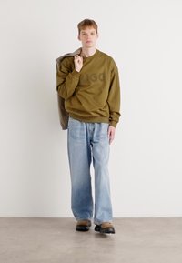 Felpa verde oliva con la scritta "HUGO", abbinata a jeans larghi blu chiaro e calzature nere. Modello che tiene una giacca esterna marrone.