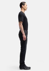 Czarny t-shirt z krótkim rękawem w połączeniu z ciemnymi jeansami i skórzanymi butami, widziany z boku, podkreślający smukły, dopasowany krój.