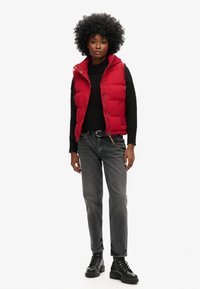 Chaleco acolchado rojo con cuello alto, suéter de punto negro, jeans rectos grises y botas militares negras con cordones. Superficies suaves y texturizadas.