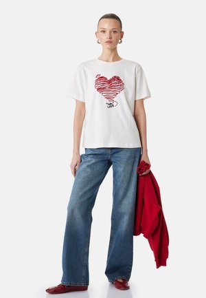 Jonge vrouw draagt een wit T-shirt met een rode hartafbeelding en de tekst "LOVE", blauwe spijkerbroek, rode schoenen, houdt in één hand een rode trui vast.