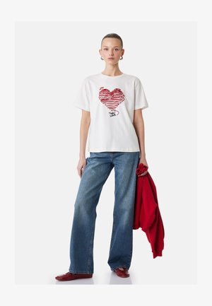 Nuori nainen pukeutunut valkoiseen t-paitaan, jossa on punainen sydän ja teksti "LOVE", siniset farkut, punaiset kengät, ja pitää toisessa kädessään punaista hupparia.