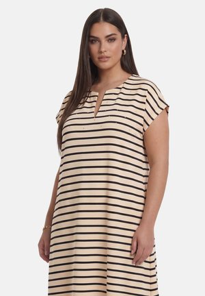 Vrouw met lang bruin haar die een beige jurk draagt met zwarte horizontale strepen en gouden oorbellen, staand tegen een witte achtergrond.