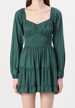 Kobieta w krótkiej, długorękawowej sukience w kolorze leśnej zieleni z marszczonym gorsetem i warstwową spódnicą, stojąca na tle jednolitego jasnego tła.