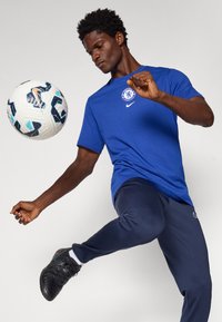 Blå Nike t-shirt med Chelsea-logo, mørke bukser, udfører fodboldtræk med en sort og hvid mønstret fodbold i luften.