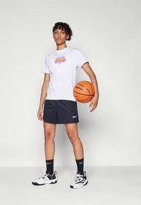 Balta marškinėliai su violetiniu grafiniu logotipu, juodos sportinės šortai, juodos Nike kojinės ir juodi bei balti sportbačiai; laiko krepšinį.