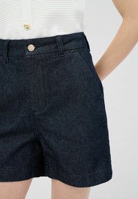 Denimshorts i mörkblått med en slät yta. Har en knappstängning, sidofickor och en rak skärning ovanför knät.
