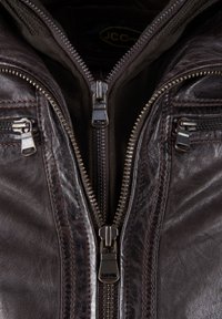 Zbliżenie na częściowo rozpiętą ciemnobrązową skórzaną kurtkę z wieloma metalowymi zamkami błyskawicznymi i widocznymi detalami ściegów.