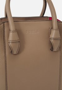 Beige nahkalaukku, jossa on jämäkkä muotoilu, kahdella lyhyellä kahvalla, ommelluilla yksityiskohdilla ja kohokuvioidulla "Furla"-logolla. Sisätila on punainen.