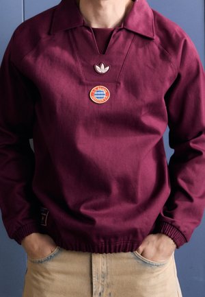 Personne portant un sweat-shirt Adidas bordeaux avec le logo du FC Bayern Munich sur la poitrine, les mains dans les poches d'un pantalon beige, devant un fond bleu.