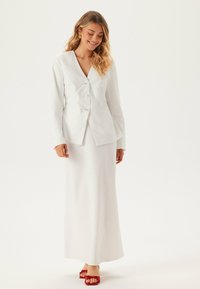 Vit kavaj med tre knappar, kombinerad med en lång vit kjol. Outfiten har en slät textur och en minimalistisk design. Röda skor accentuerar looken.