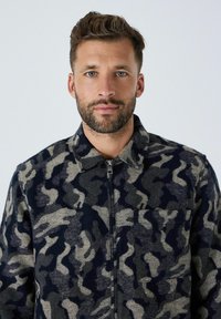 Man met een baard en golvend haar, gekleed in een zwart-grijs camouflagedessin zip-up jack tegen een effen lichte achtergrond.