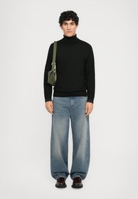 Schwarzer Rollkragenpullover, lockere hellblaue Jeans und dunkelbraune Schuhe. Das Modell trägt eine kleine olivgrüne Umhängetasche.