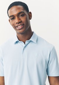 Šviesiai mėlynas polo marškinėliai su minkšta tekstūra, turintys trumpą apykaklę ir mažą siuvinėtą logotipą kairėje pusėje. Dvi mygtukų užsegimo apykaklėje.