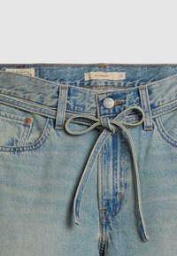 Jean en denim bleu clair avec un bouton et une ceinture en denim nouée à la taille, montrant les poches avant et l’étiquette de taille 27 XL coupe droite.