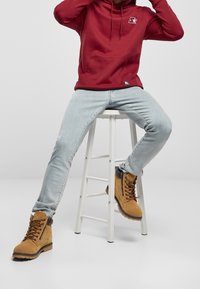 Hoodie rouge avec une poche frontale, jeans bleu clair et bottes marron clair avec accents noirs. Le hoodie présente un logo sur la poitrine.