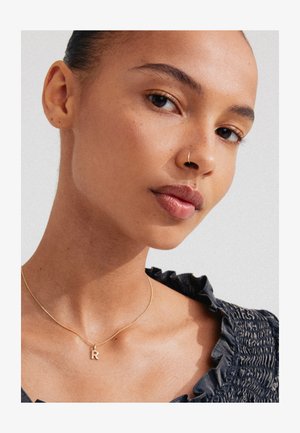 Guld halskæde med et bogstav "R" vedhæng båret på en glat hals over en mørk, rynket stoftop; hudtonen er varm med subtil tekstur.