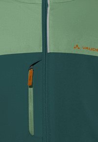 Giacca da esterno verde e turchese con un pannello verde chiaro a contrasto, dotata di una tasca con zip e un tirante arancione, oltre al logo del marchio.