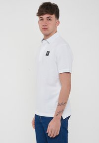 Polo shirt bianca con colletto classico, maniche corte, una toppa nera con logo sul petto e un orlo diritto. Adatta per un abbigliamento casual.
