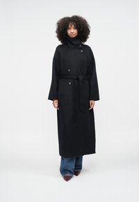 Lang sort frakke lavet af uld, med høj krave, dobbeltknappet og bånd i taljen, kombineret med vidde jeans og spidse sko.