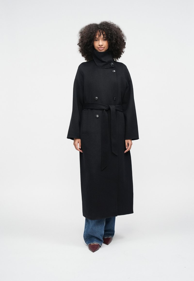 Lang sort frakke lavet af uld, med høj krave, dobbeltknappet og bånd i taljen, kombineret med vidde jeans og spidse sko.