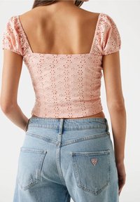 Kvinde iført en lys pink broderet cropped top med pufærmer og højtaljede lyseblå denim jeans med et synligt mærke-logo.