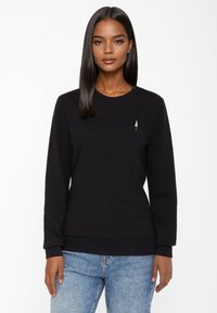 Zwarte sweater met lange mouwen, ronde halslijn en een klein geborduurd boomlogo op de linkerkant van de borst. Gecombineerd met lichtblauwe jeans.