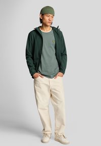 Mørkegrøn letvægtsjakke over en grå t-shirt med et broderet logo, kridtfarvede bukser og beige sneakers. Rund ribbet hue.