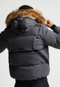 Hall sinine pusa, kus on karvane kapuuts. Omab reguleeritavaid rihmasid ja seljal punase vahtralehe logoga plaastrit.