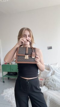 Barna, geometriai mintájú kézitáska felső fogantyúval, aranyszínű fémrészletekkel és láncos díszítéssel. Strukturált, dobozszerű formája van és texturált felületű.