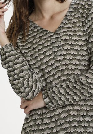 Kvinde iført en grøn mønstret bluse med lange ærmer og muslingeformede designs, holder sit hår med den ene hånd og har korslagte arme.