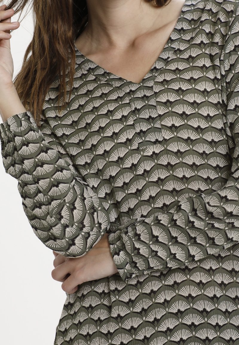 Femme portant un haut vert à manches longues avec des motifs en forme de coquilles, tenant ses cheveux d'une main et bras croisés.