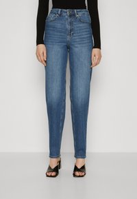 Τζιν ψηλής μέσης σε μπλε απόχρωση με ίσια γραμμή, φτιαγμένα από τζιν ύφασμα. Διαθέτουν πέντε τσέπες και ραφές, συνδυασμένα με μαύρες σαμπάνιες με τακούνι.