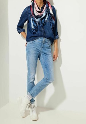 Femme portant une chemise en jean retroussée, un jean bleu clair, une écharpe colorée et des baskets blanches, debout contre un mur blanc.