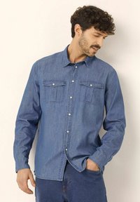 Camicia in denim di blu medio, con chiusura a bottoni sul davanti, due tasche sul petto e colletto classico. Tessuto liscio con maniche lunghe.
