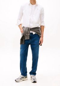 Camicia bianca a bottoni indossata sopra un sweatshirt grigio legato in vita, jeans blu e sneaker grigie con dettagli neri e suole bianche.