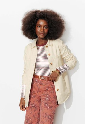 Femme aux cheveux bouclés volumineux portant une veste crème boutonnée, un chemisier rayé à manches longues, un pantalon à fleurs et des bracelets dorés, posant devant un fond blanc.