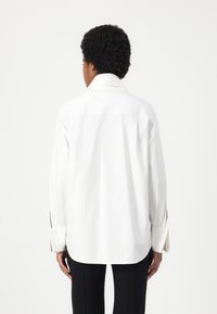 White shirt featuring a pointed collar, single-button cuffs, and a relaxed fit with a rounded hem, worn with black trousers.