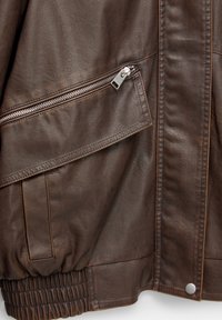 Gros plan sur une veste en cuir marron avec une poche zippée en biais, une couture latérale verticale, une ceinture élastique et un bouton-pression métallique à l’ourlet.