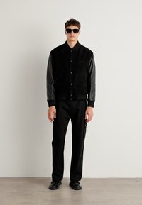 Black suede bomber jacket with leather sleeves, featuring snap buttons and a minimalist design. Black pants and sunglasses complete the look.
