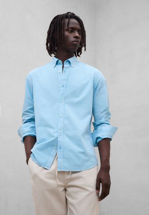 Mand med mørke dreadlocks iført en lyseblå skjorte med knapper og beige bukser, stående foran en ensfarvet grå baggrund.