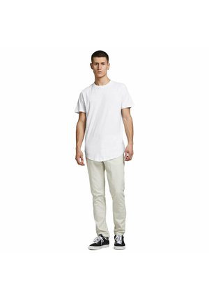 Jonge man staand, dragend een eenvoudig wit T-shirt, beige broek en zwarte sneakers met witte veters op een witte achtergrond.