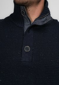 Marineblauer Strickpullover mit strukturiertem Muster, ausgestattet mit einem Reißverschluss und zwei Knöpfen am Kragen, sowie einer kontrastierenden, denimähnlichen Innenseite.