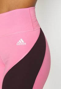 Růžové a černé legíny s vysokým pasem, s hladkou texturou, obsahující bílé logo Adidas a kontrastní zakřivený panelový design.