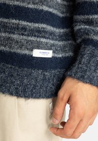 Navyblauwe gebreide trui met lichtgrijze strepen, fuzzy textuur, en label met 'ELEMENT CO. SINCE 1992'. Beige stof zichtbaar eronder.
