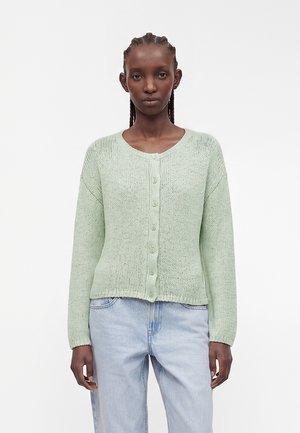 Frau mit geflochtenem Haar, die eine hellgrüne, geknöpfte Strickjacke und hellblaue Jeans trägt, steht vor einem schlichten weißen Hintergrund.