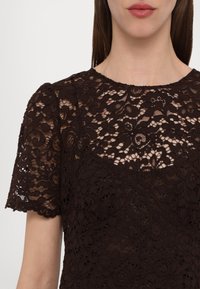 Femme portant un haut en dentelle noire à manches courtes avec des motifs floraux et un col rond, dévoilant le haut de la poitrine et les épaules.