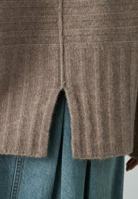 Jasnobrązowy sweter z prążkowanego dzianiny z wysokim rozcięciem na dolnej części pleców, w zestawieniu z niebieską spódnicą dżinsową. Miękka tekstura i luźny krój.