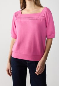 Žena v jasně růžovém topu s krajkovým čtvercovým výstřihem a rukávy do lokte, doplněná tmavě modrými džíny na jednolitém pozadí.