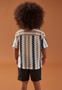 Kurzärmeliges Hemd mit Zickzackmuster in Creme, Braun und Navy, kombiniert mit dunklen Shorts. Strukturiertes Gewebe, lockere Passform.